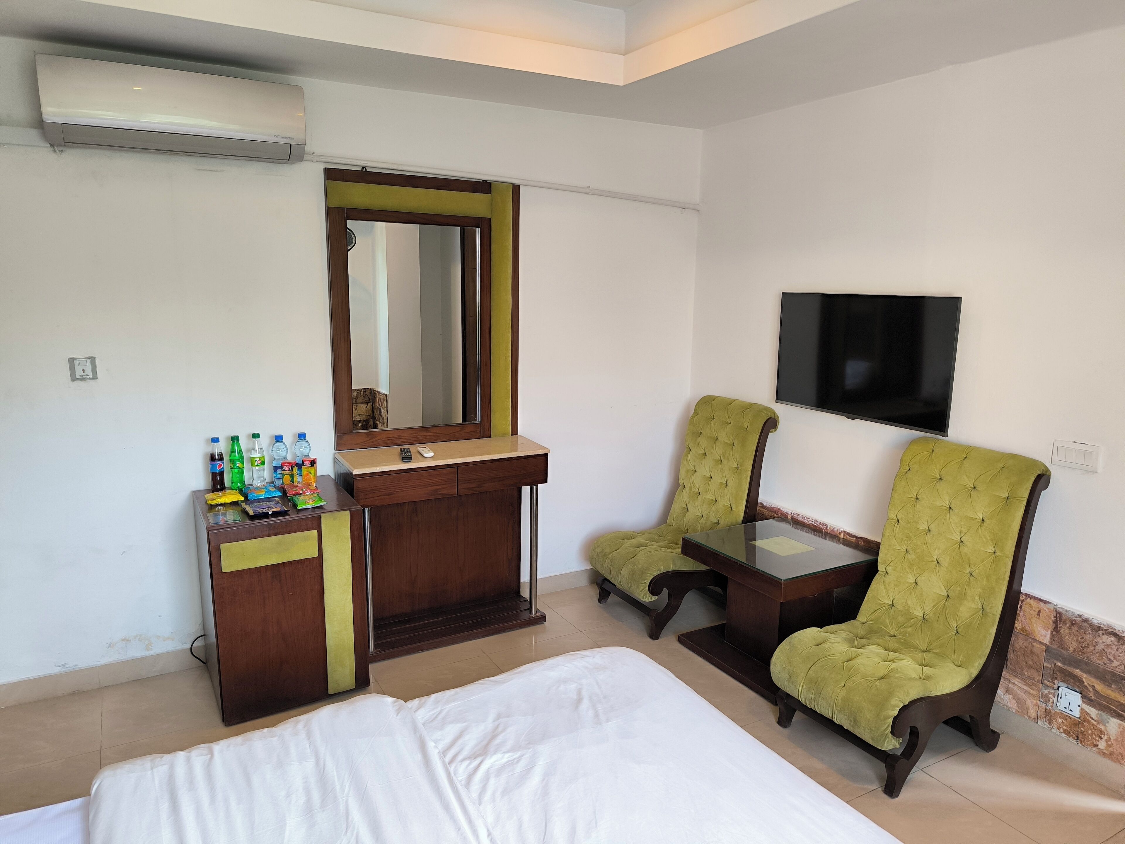 Habitación de hotel con una cama, un tocador con espejo, dos sillones tapizados en verde, un televisor de pantalla plana y una nevera con bebidas.