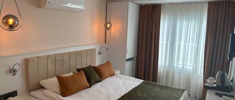A hotel room with a bed, bedside table, lamp, air conditioning unit, curtains, and a window.