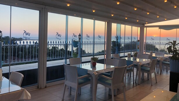 En terrasse med glasborde og stole, der giver udsigt over et smukt hav- og bjerglandskab i solnedgang.