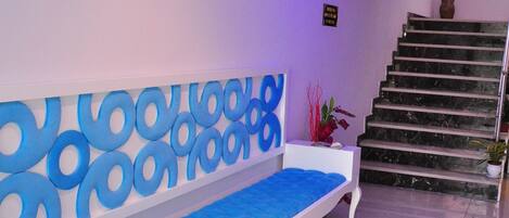 Un área de recepción moderna con un banco blanco, una pared con diseño azulado, una escalera y una planta en maceta.