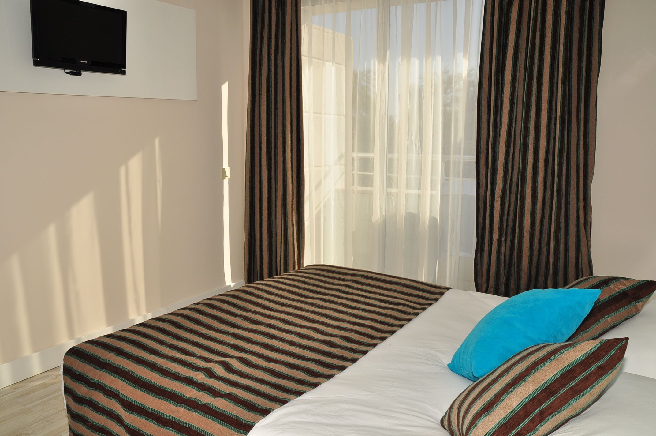 A hotel room with a bed, striped curtains, a television on the wall, and a window with sheer curtains.