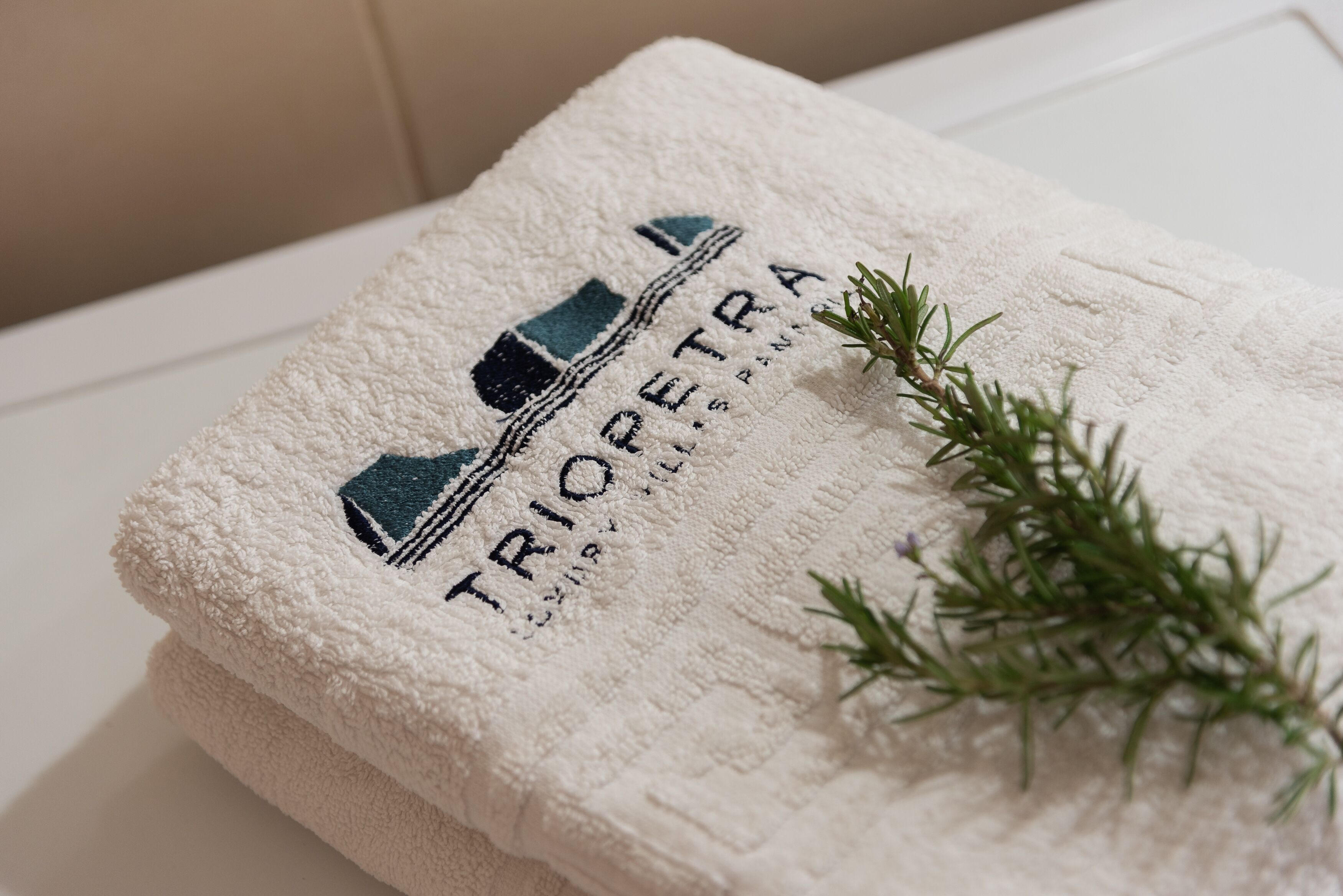 A white towel with embroidered text and a logo, placed on a white surface with a sprig of rosemary.