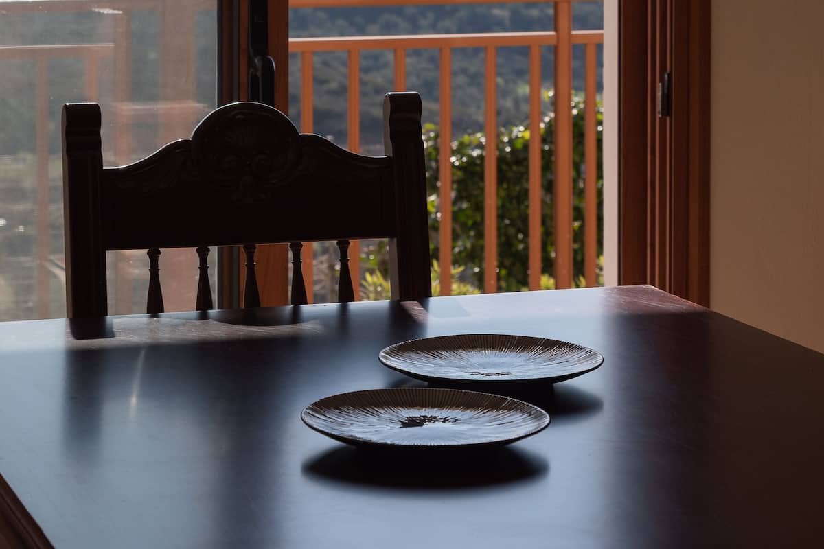 Una mesa de comedor con dos platos y una silla, con vistas a un paisaje montañoso y una masa de agua a través de la barandilla de un balcón.
