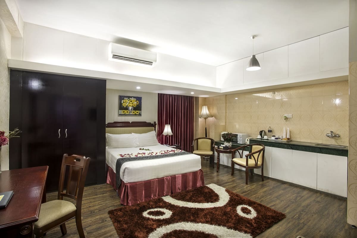 Habitación de hotel con cama, escritorio, silla, mesita, lámpara y kitchenette.