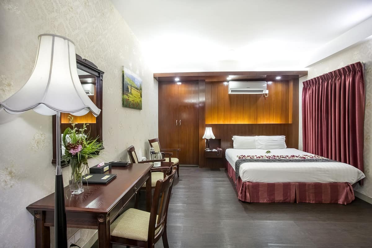 Habitación de hotel con escritorio de madera, cama con cubrecolchón rojo, mesita de noche, lámpara y unidad de aire acondicionado.
