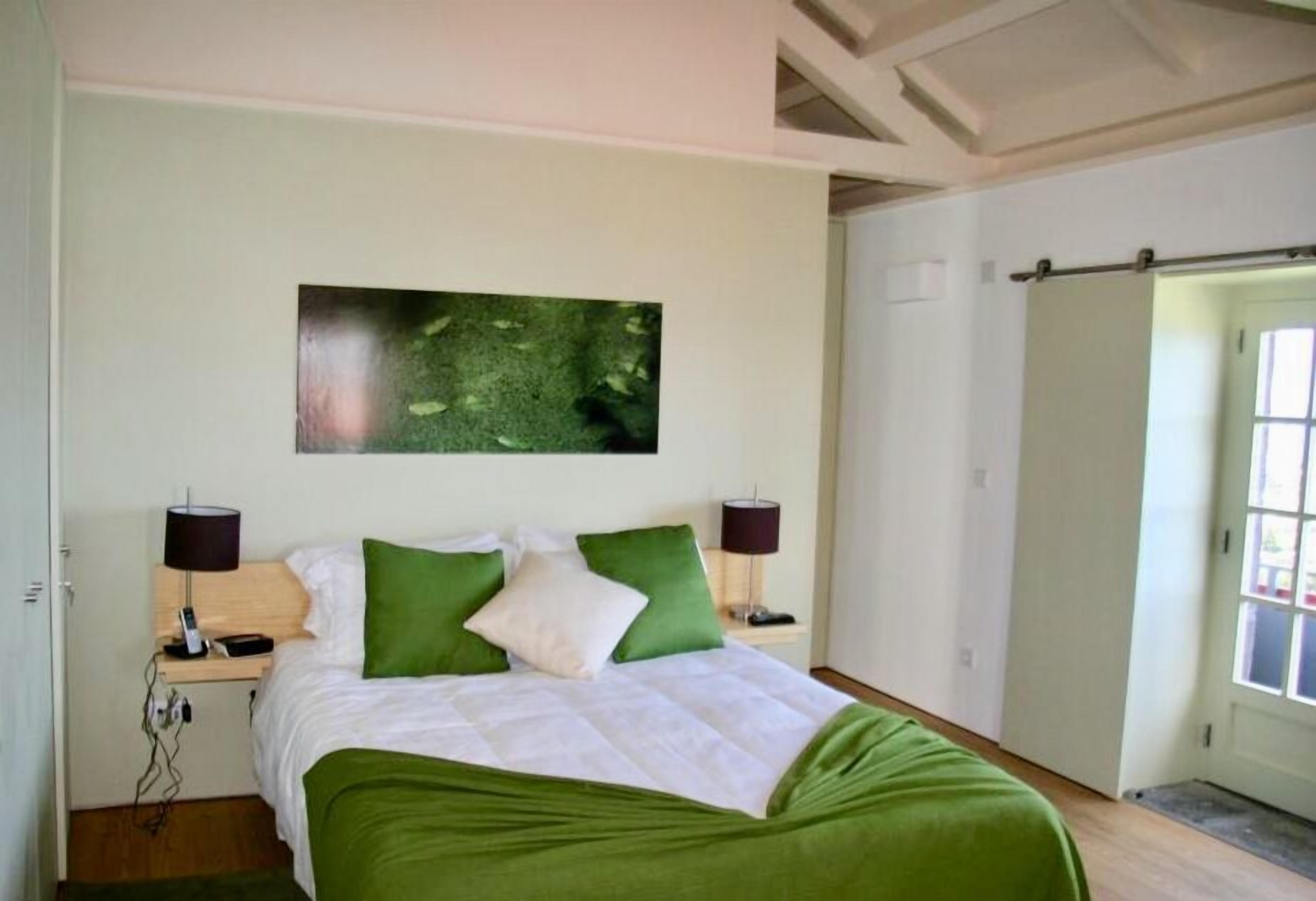 A bedroom with a bed, green pillows, two bedside lamps, a nightstand, a wall-mounted artwork, and a sliding door.
