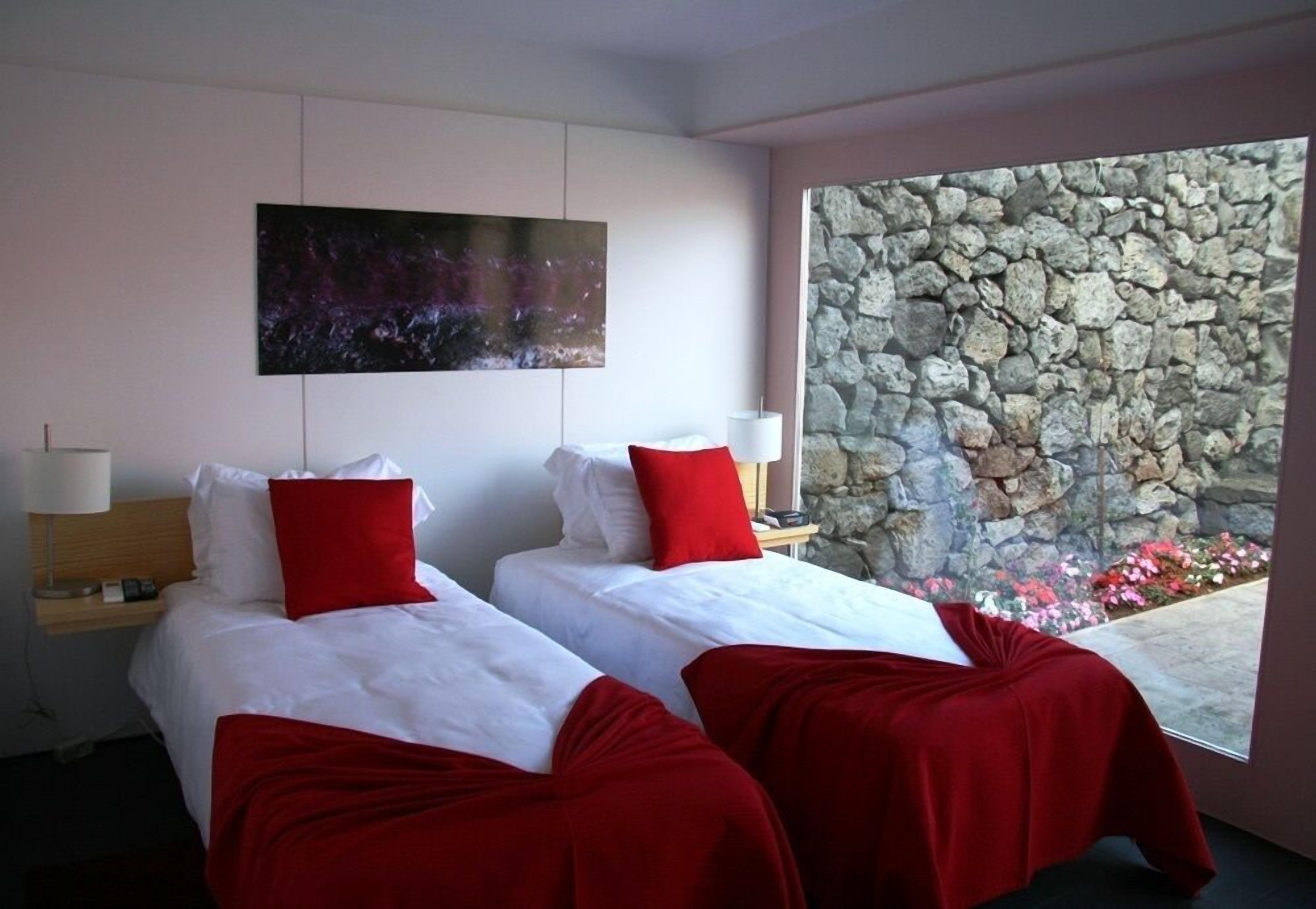 Une chambre d’hôtel avec deux lits, une literie rouge et blanche, un grand tableau au mur et un mur de pierre orné de fleurs à l’extérieur.