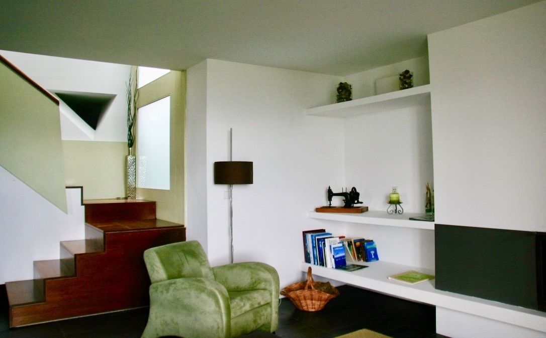 A modern living room with a staircase, a green armchair, a bookshelf, and a flat-screen TV.