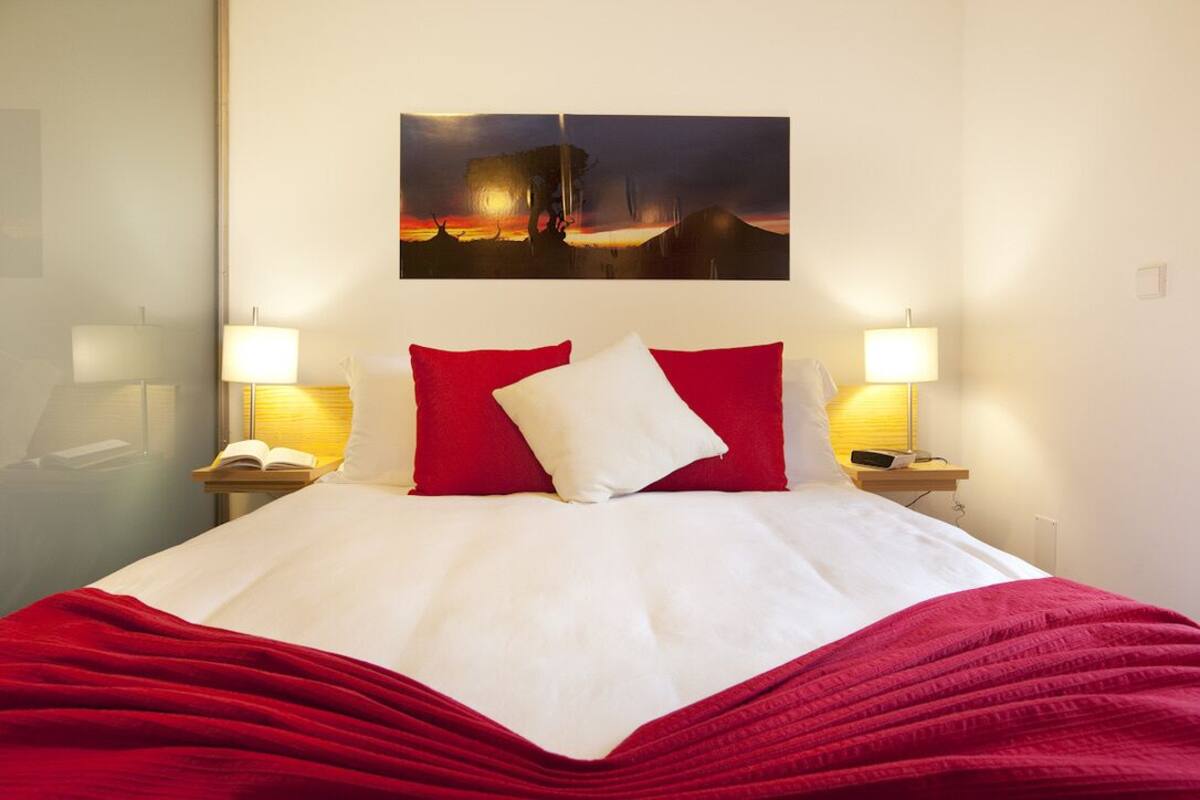 Une chambre d’hôtel avec un lit, deux lampes de chevet, une table de chevet avec un livre et un téléphone, et un tableau au mur.