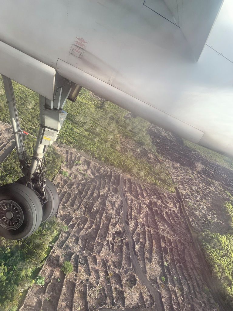 An aircraft wing with a visible landing gear and a ground below, featuring a patterned landscape with vegetation.