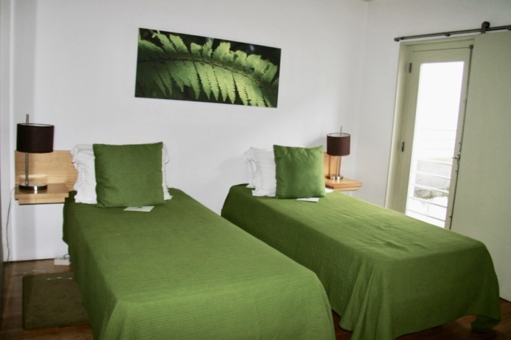 Une chambre avec deux lits simples, chacun recouvert d'une couverture verte et agrémenté d'oreillers assortis. On y trouve un tableau représentant une fougère accroché au mur, une table de chevet avec une lampe, et une porte sur le côté droit.