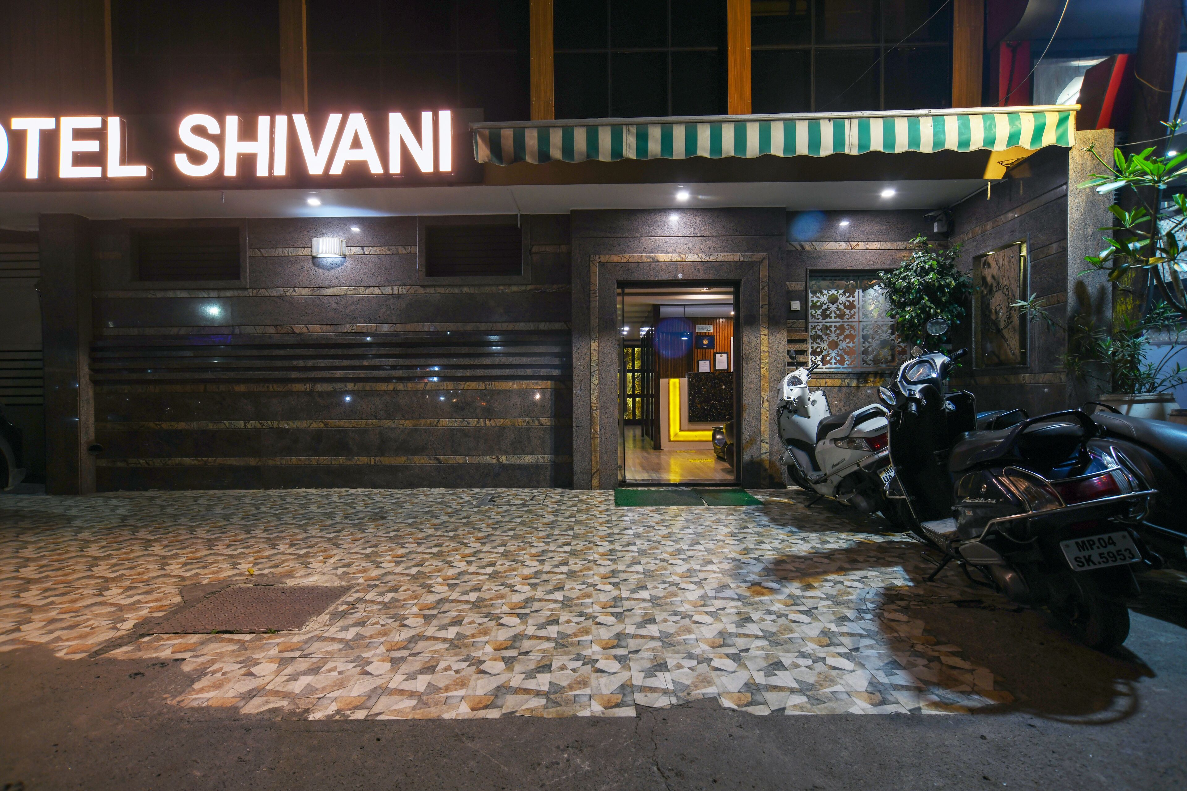 Exterior del Hotel Shivani con un acceso empedrado, una motocicleta estacionada afuera y un letrero de neón sobre la entrada.
