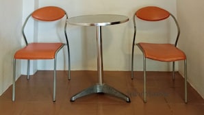 Una mesa pequeña y redonda con base de metal y dos sillas naranjas de estructura metálica.