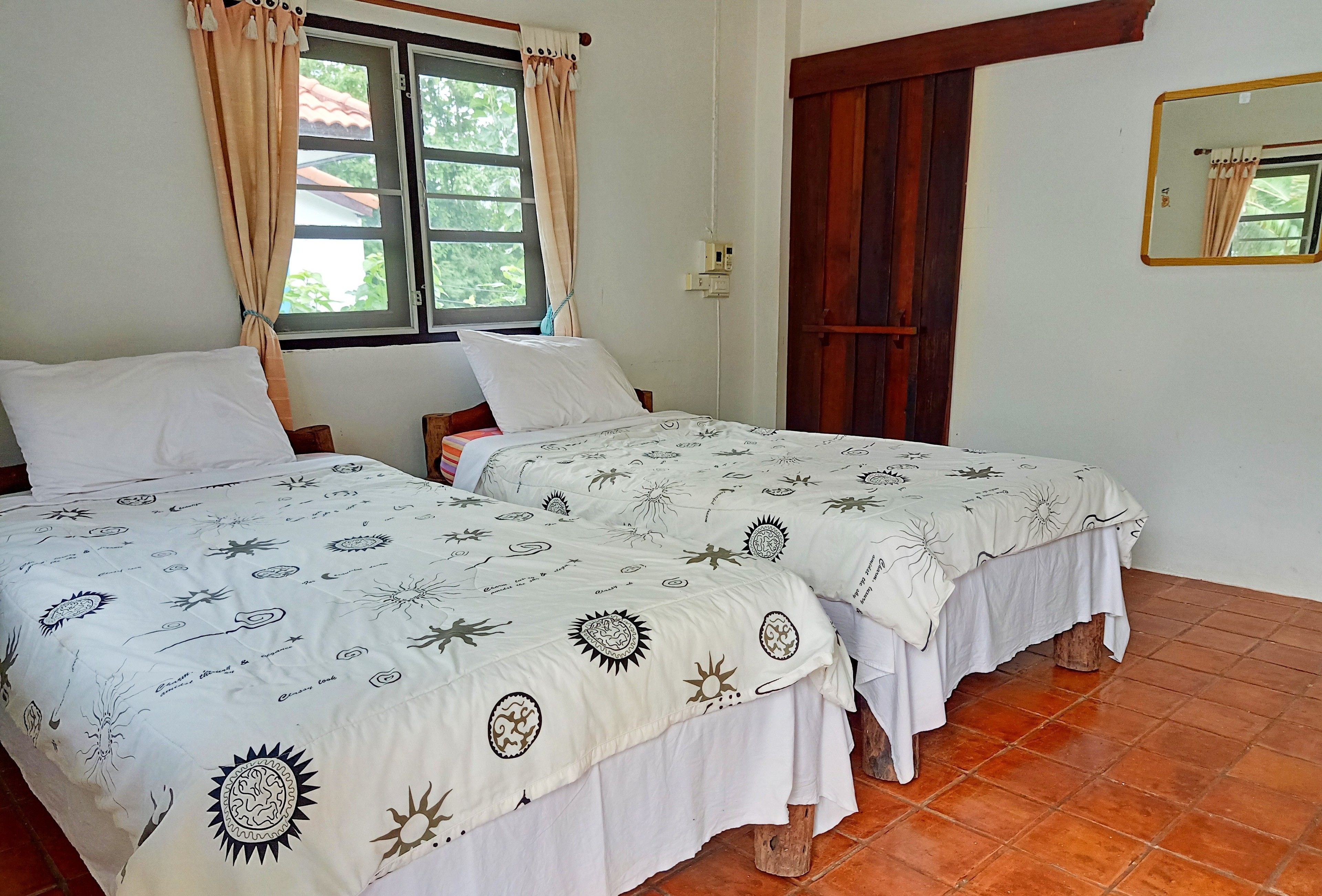 A room with two beds, each with a sun-patterned bedspread, a window with curtains, a wooden door, and a mirror on the wall.