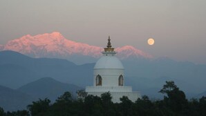 A white building with a dome and a spire, surrounded by trees and mountains in the background.