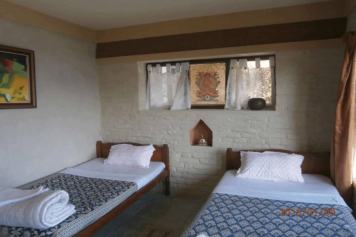 Une chambre avec deux lits, une fenêtre avec des rideaux, une petite étagère ornée d’un objet décoratif et un tableau accroché au mur.