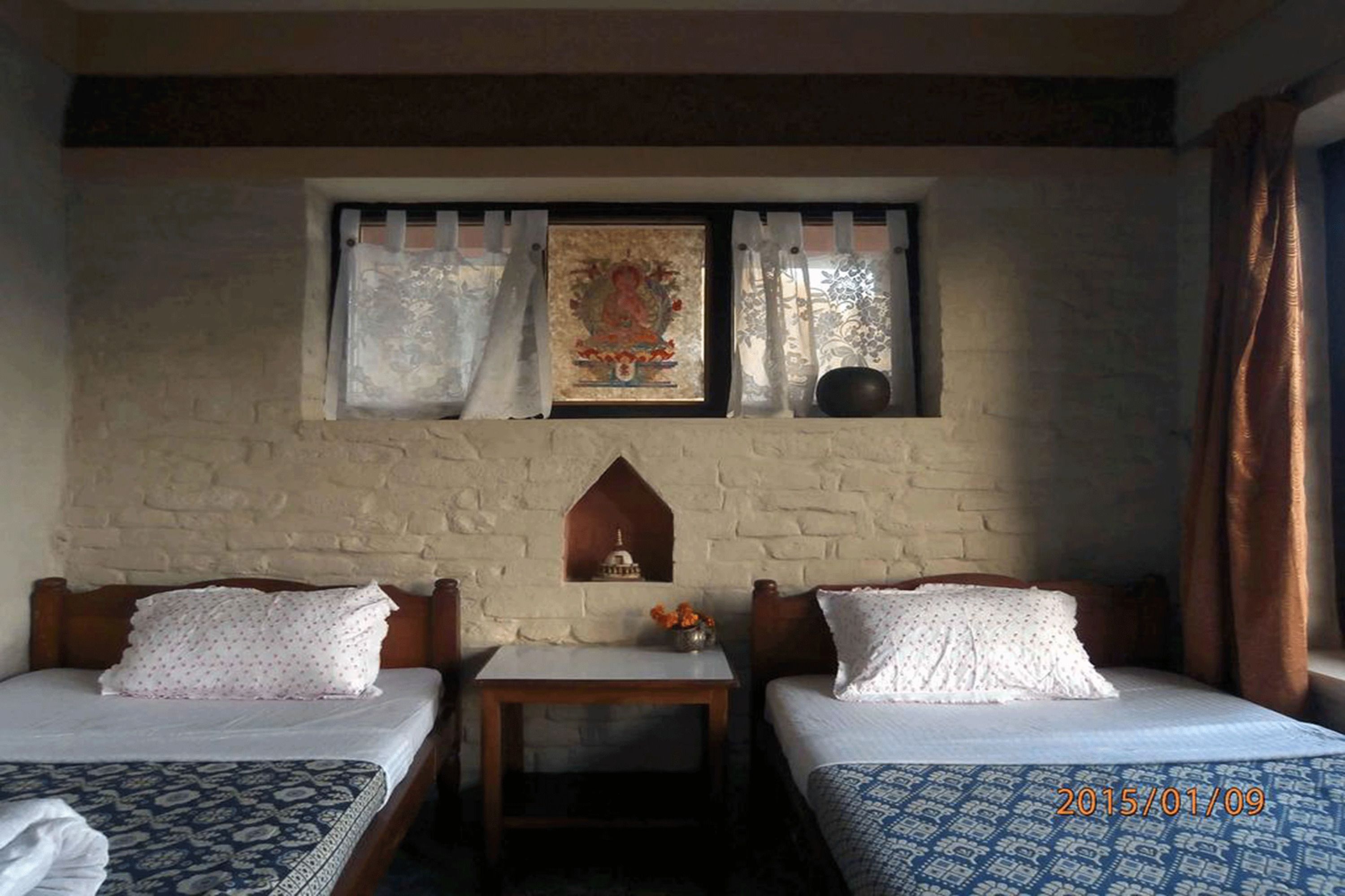 A room with two beds, a small table, a decorative wall hanging, and a window with curtains.