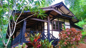 A traditional wooden house with a balcony, surrounded by lush greenery and a brick wall.