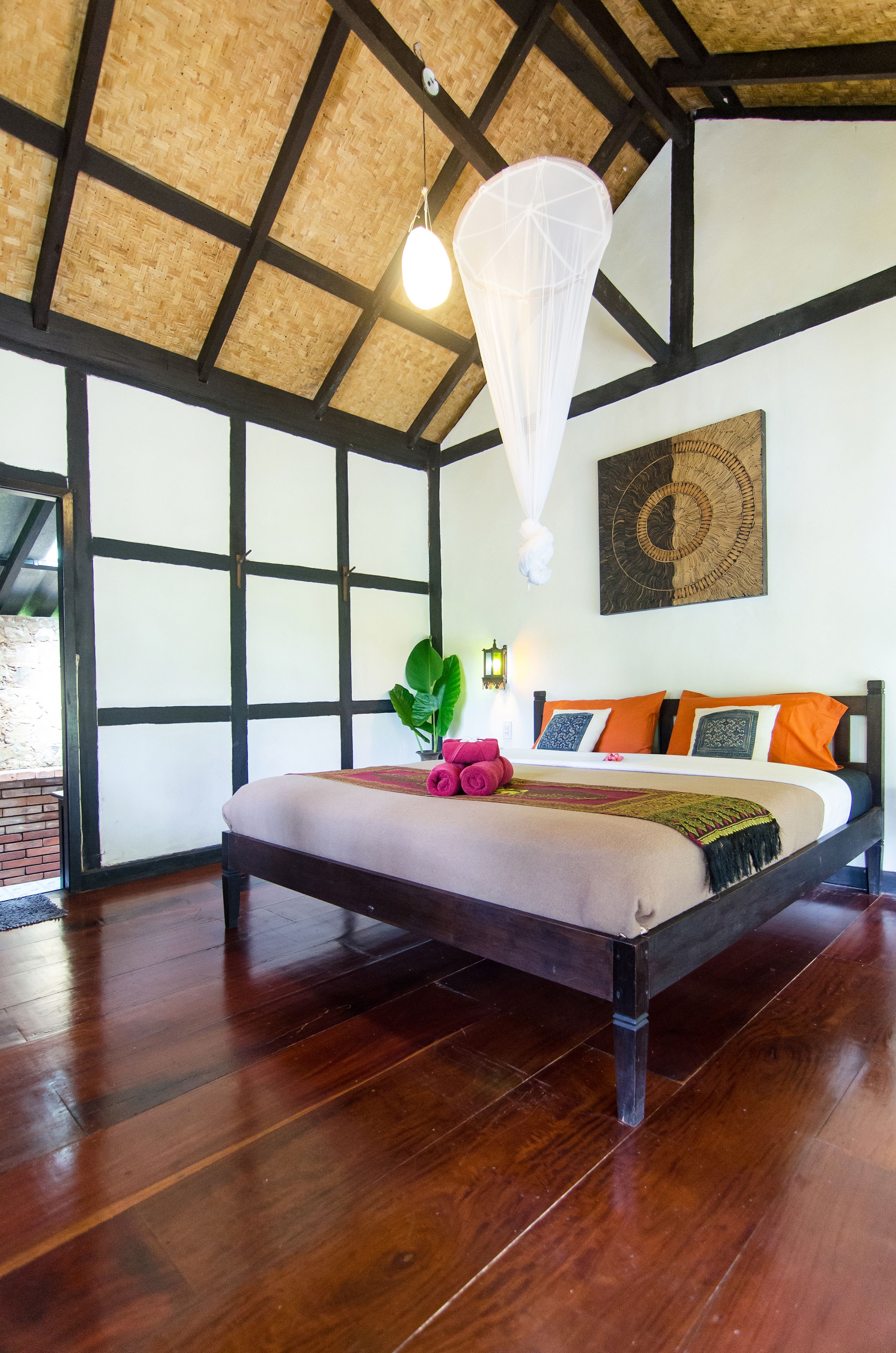 A bedroom with a wooden ceiling, a bed with orange and black pillows, and a large window.
