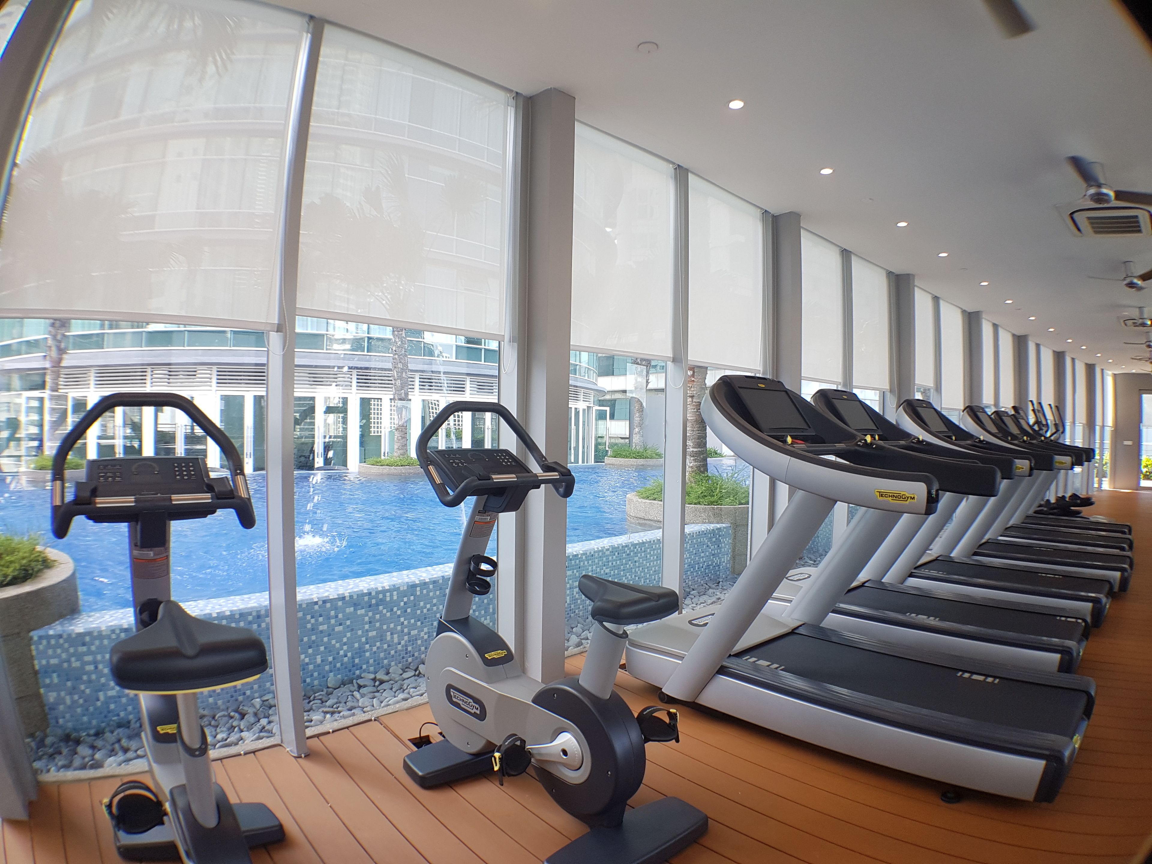Une salle de sport équipée de tapis de course et d’appareils elliptiques, dotée de grandes fenêtres et offrant une vue sur un immeuble et une piscine.