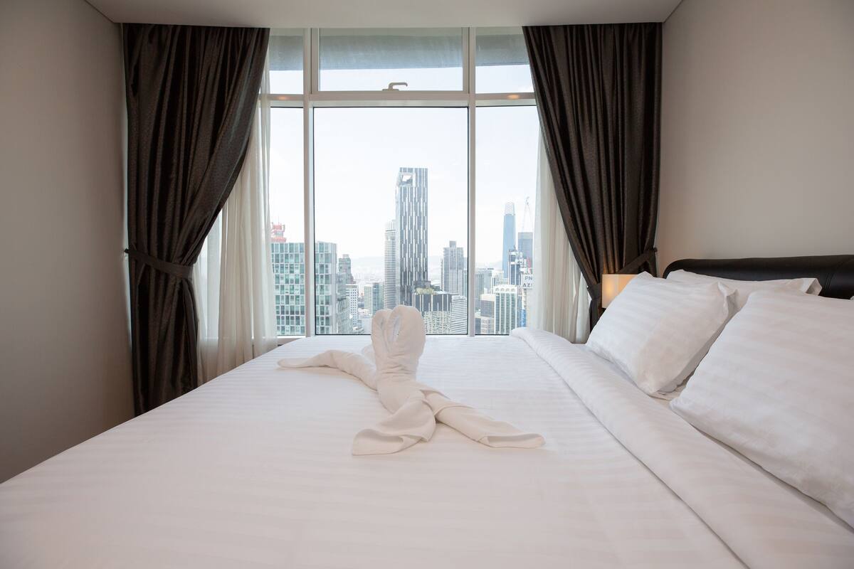 A hotel room with a large bed, white linens, and a view of a cityscape through floor-to-ceiling windows.