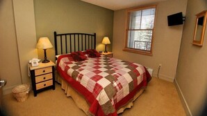 A bedroom with a bed, two bedside tables with lamps, a window with blinds, and a wall-mounted TV.