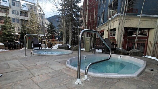 A pool area with two hot tubs, stone flooring, and surrounding buildings.