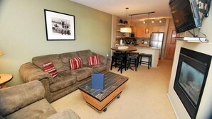 A living room with a sofa, coffee table, and fireplace. There is a kitchen area with a dining table and chairs.