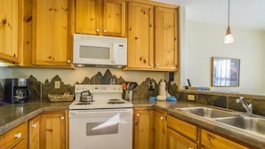 A kitchen with wooden cabinets, a white stove, a microwave, a granite countertop, and a stainless steel sink. - Snake River Village 2 Bedroom Apartment by Key to the Rockies (Keystone)