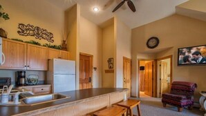 Eine Küche mit Holzausstattung, einem Kühlschrank, einer Spüle und einer Bar mit Hockern. Es gibt einen Deckenventilator, eine Uhr an der Wand und einen Sessel im Wohnbereich.
