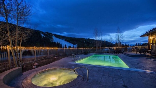 A heated outdoor pool area with a hot tub, surrounded by a wooden deck and a fence with string lights.