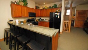 A kitchen with wooden cabinets, a granite countertop, a black refrigerator, and a microwave. There are three bar stools and a sink.