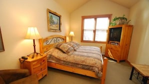 A bedroom with a wooden bed, two bedside lamps, a wooden nightstand, a wooden dresser with a television, a chair, and a window with blinds.