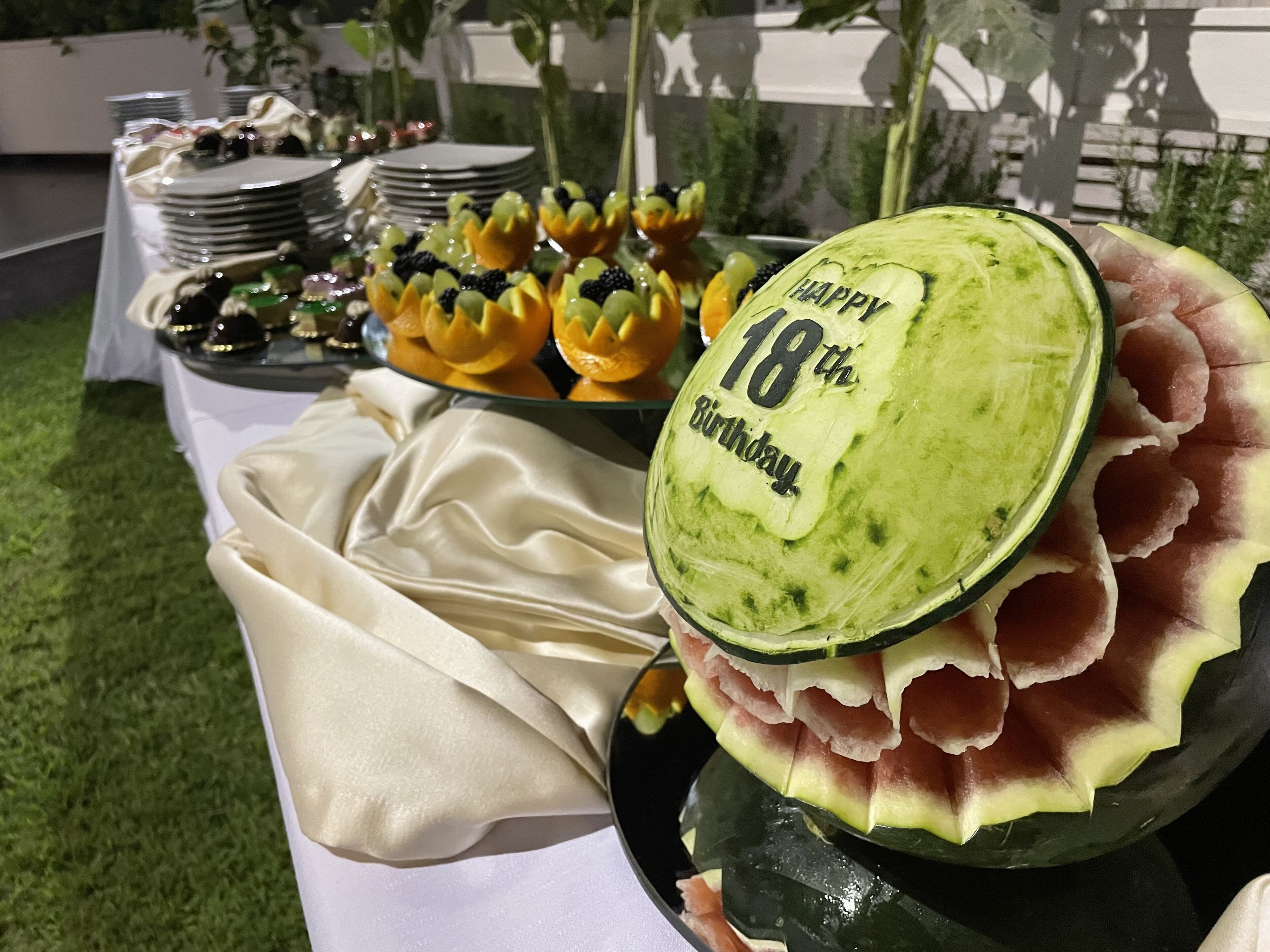 Une table décorée d’un centre de table composé d’une pastèque et portant l’inscription « Joyeux 18e anniversaire », ainsi que de diverses compositions de fruits.