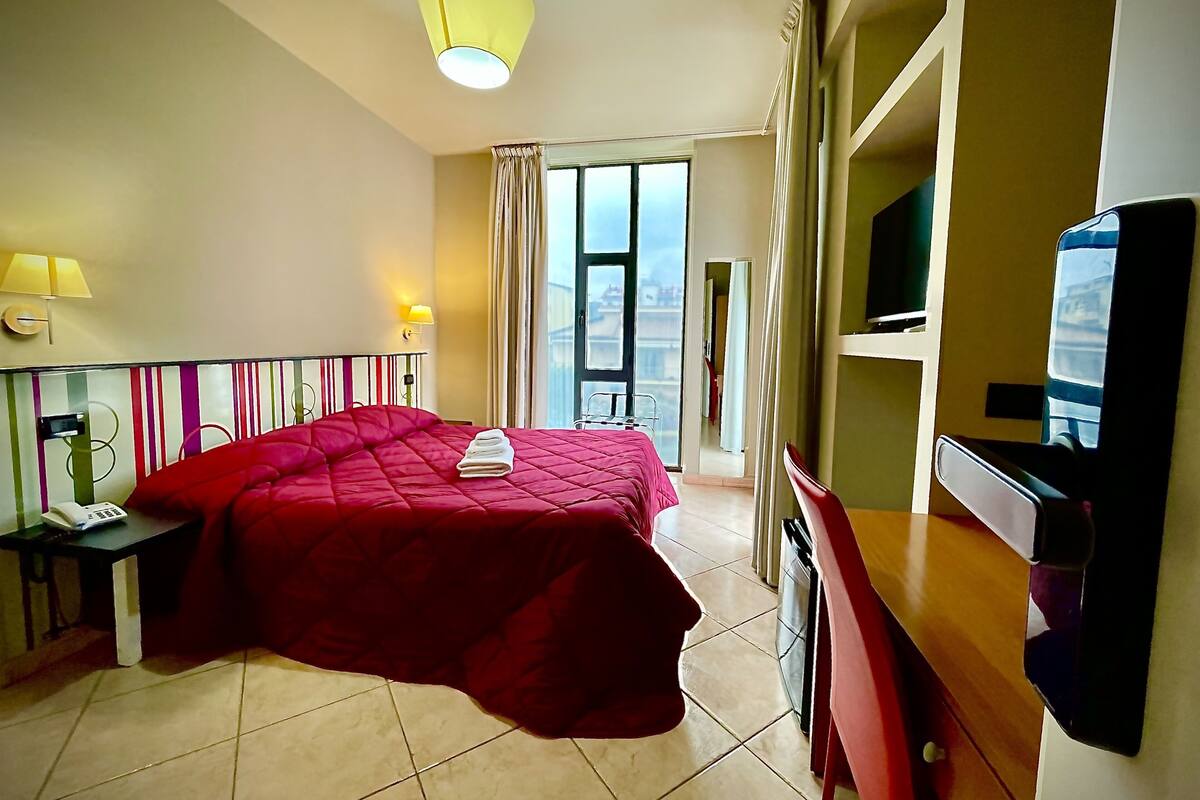 A hotel room with a large bed, a television, a desk with a telephone, and a window with curtains.