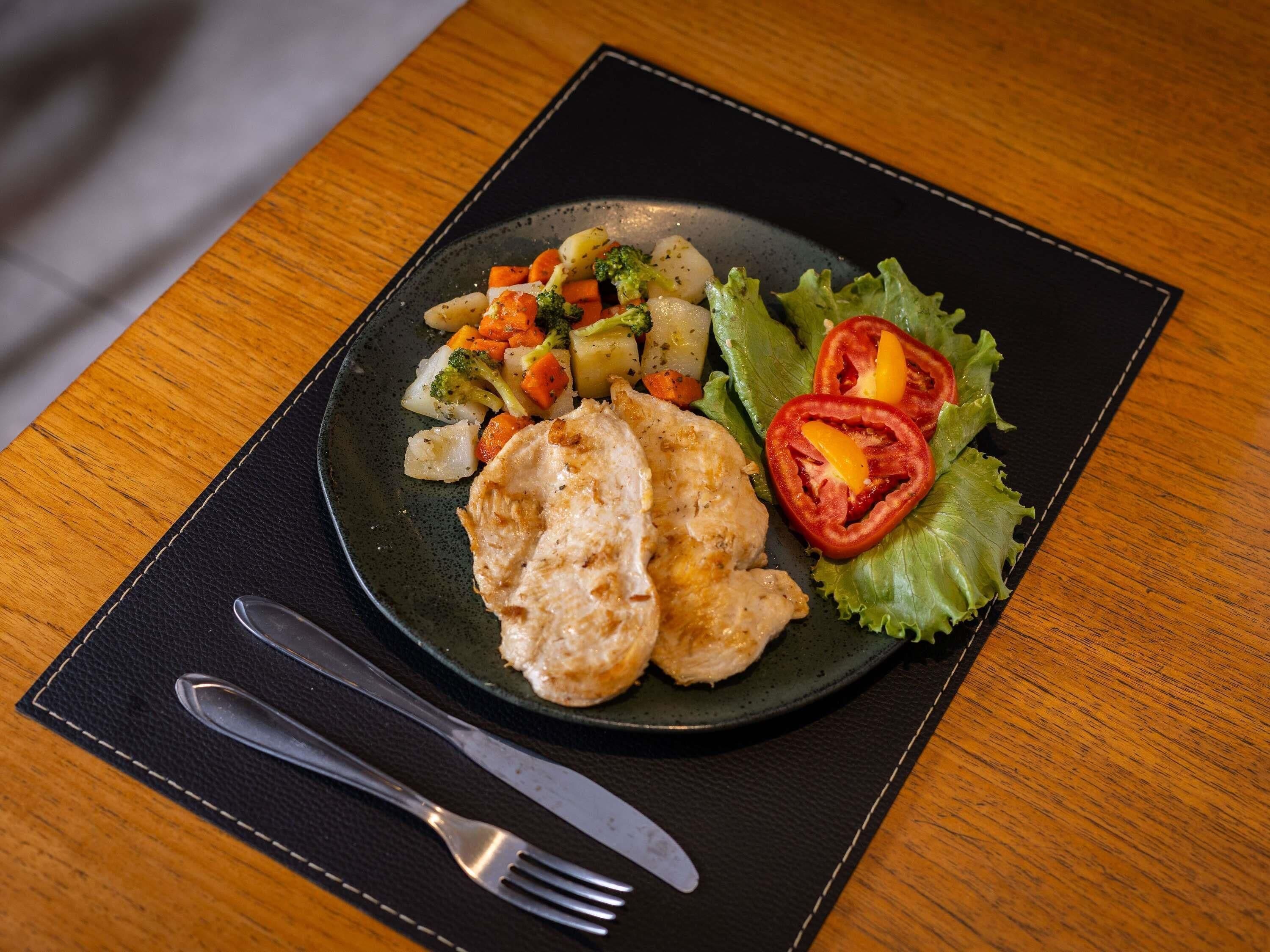 A plate of food with vegetables and a piece of chicken on a black plate, placed on a black placemat.