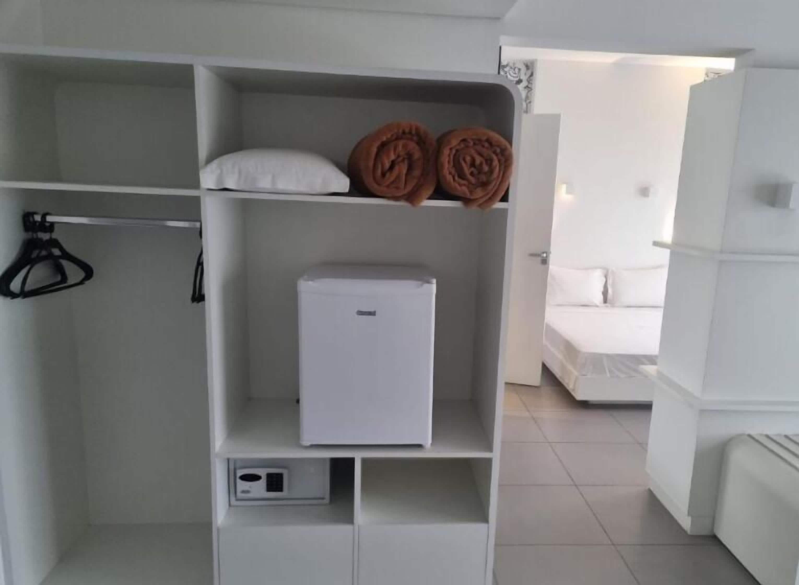 A white wardrobe with a small appliance, a hanging rack, and a bed in the background.
