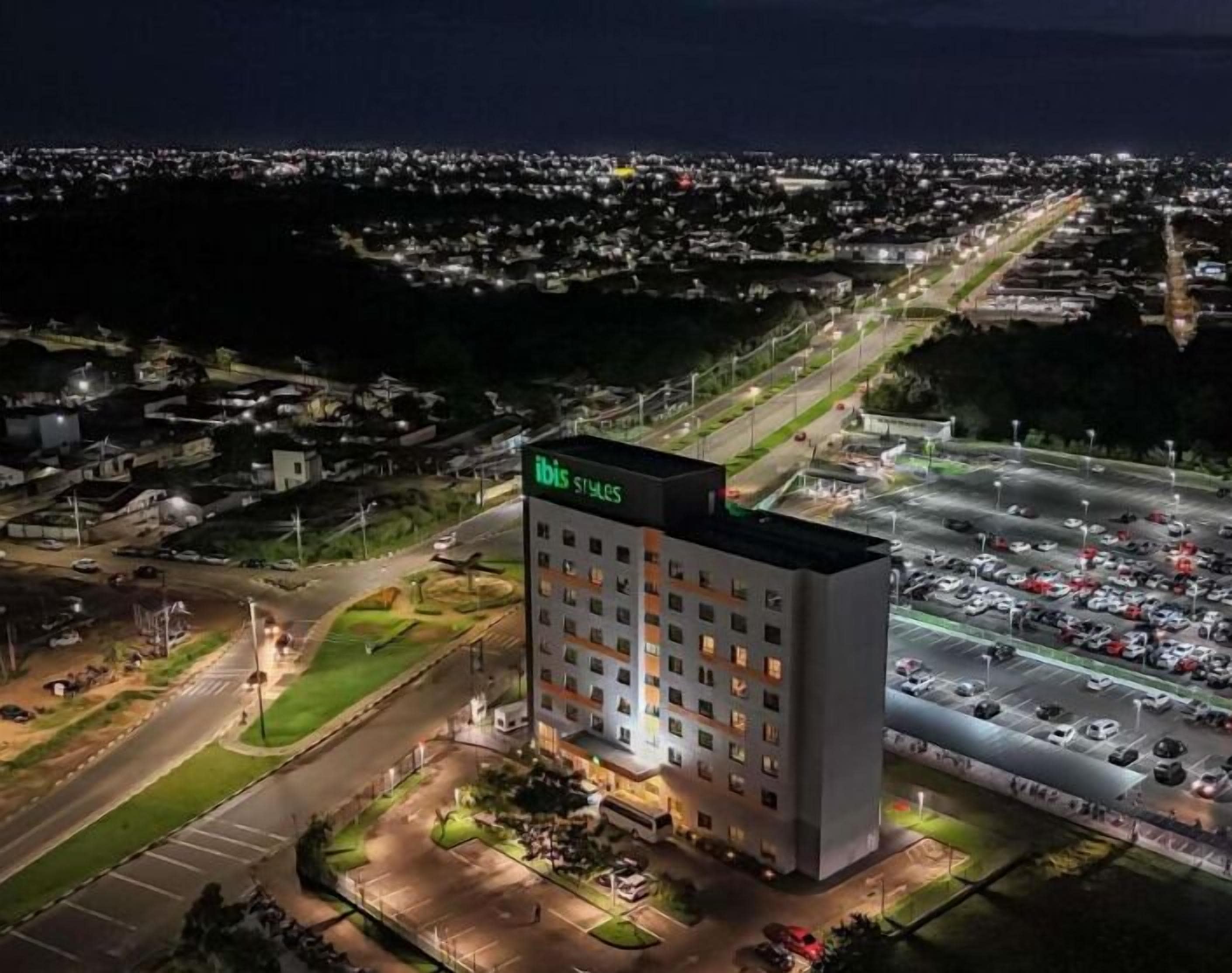 Un hotel de varios pisos con un letrero en la azotea, rodeado por un estacionamiento y una calle.