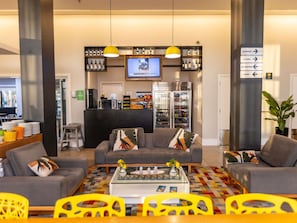 Eine moderne Lobby mit Sitzgelegenheiten, einem Fernseher, einem Kühlschrank und einem Schild, das Annehmlichkeiten wie Aufzüge und eine Lounge anzeigt.