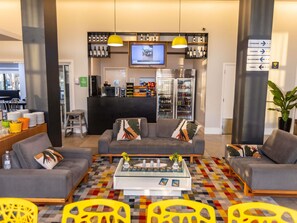 Un hall de hotel con sofás, una mesa de centro, un televisor, un refrigerador y un letrero que indica servicios como ascensores, un salón y una sala de reuniones.