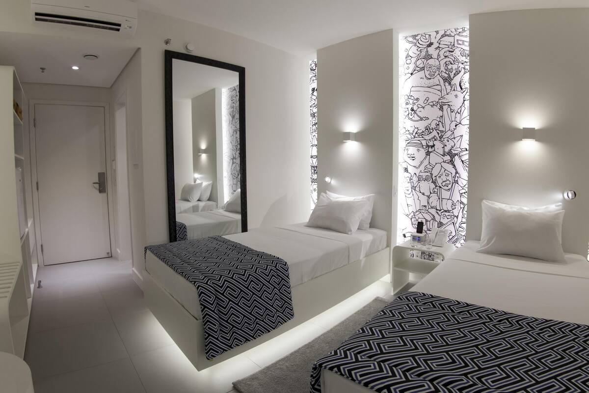 Ett modernt hotellrum med en stor säng, en mindre säng, en spegel och en vägg med ett svartvitt mönster.