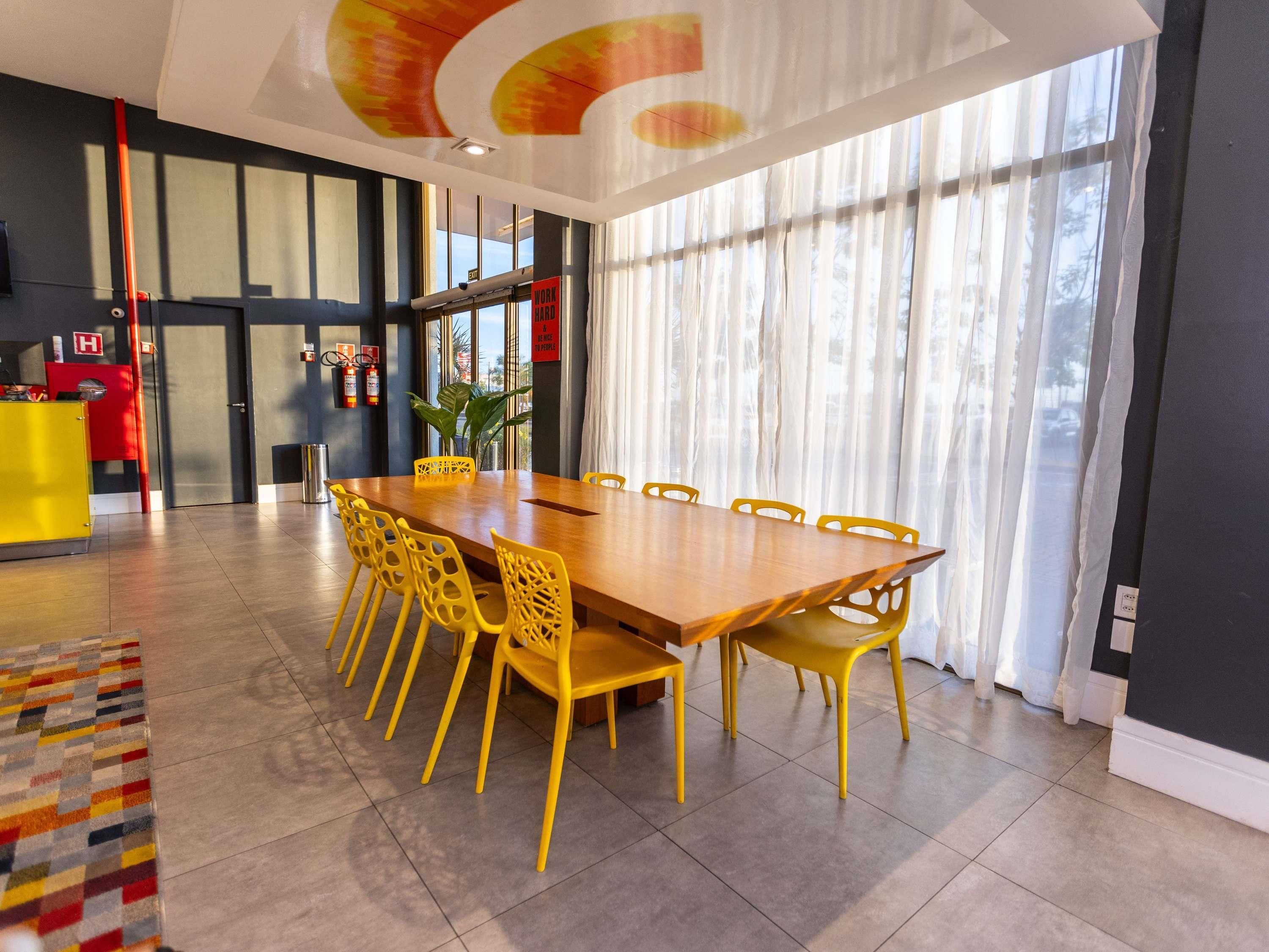 A modern hotel room with a large wooden dining table, yellow chairs, a red minibar, and a large window with curtains.