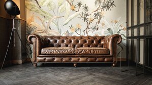 A brown leather sofa in a room with a herringbone-patterned wooden floor, a large window with blinds, and a wall mural of tropical foliage. - Hotel Rosengarten  (Zweibruecken)