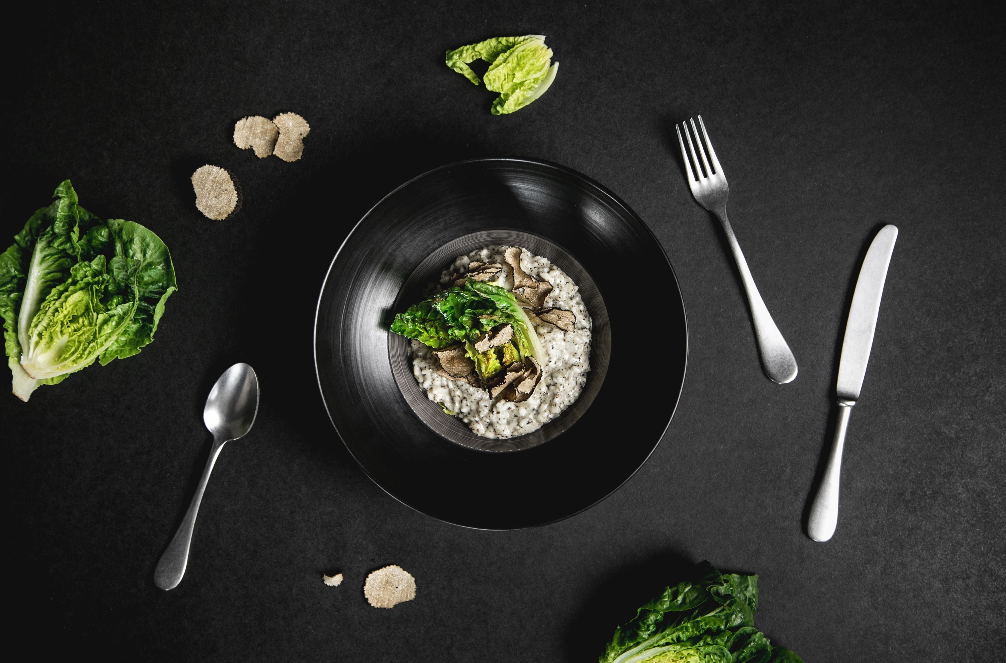 Un plato negro con ensalada sobre una superficie oscura, acompañado de un tenedor, cuchillo y cuchara.