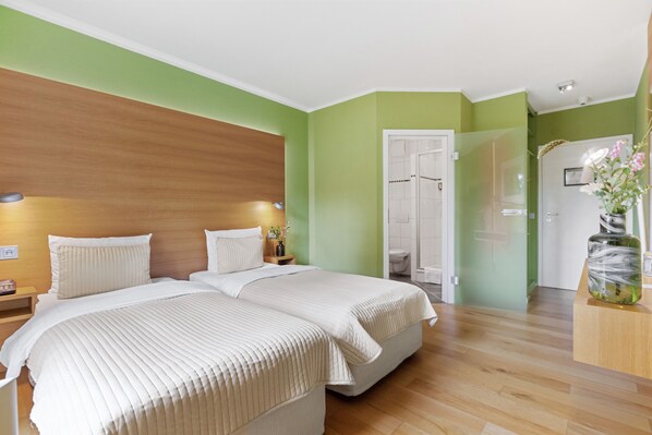 A hotel room with two beds, a wooden headboard, green walls, a wooden floor, a bathroom with a glass door, and a vase with flowers on a wooden table. - Hotel Rosengarten (Zweibruecken)