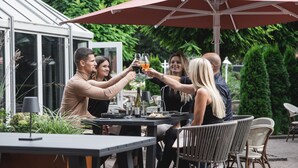 A group of people toasting with drinks at an outdoor table with a large umbrella, surrounded by greenery and a greenhouse structure. - Hotel Rosengarten  (Zweibruecken)