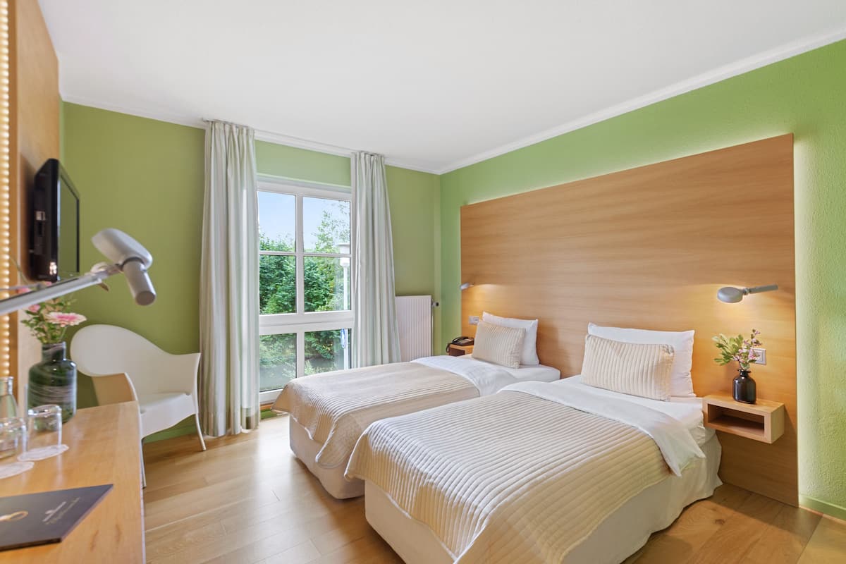 Ein Hotelzimmer mit zwei Betten, einem Schreibtisch mit Lampe, einem Sessel, einem Fenster mit Vorhängen und einem Holz-Kopfteil.