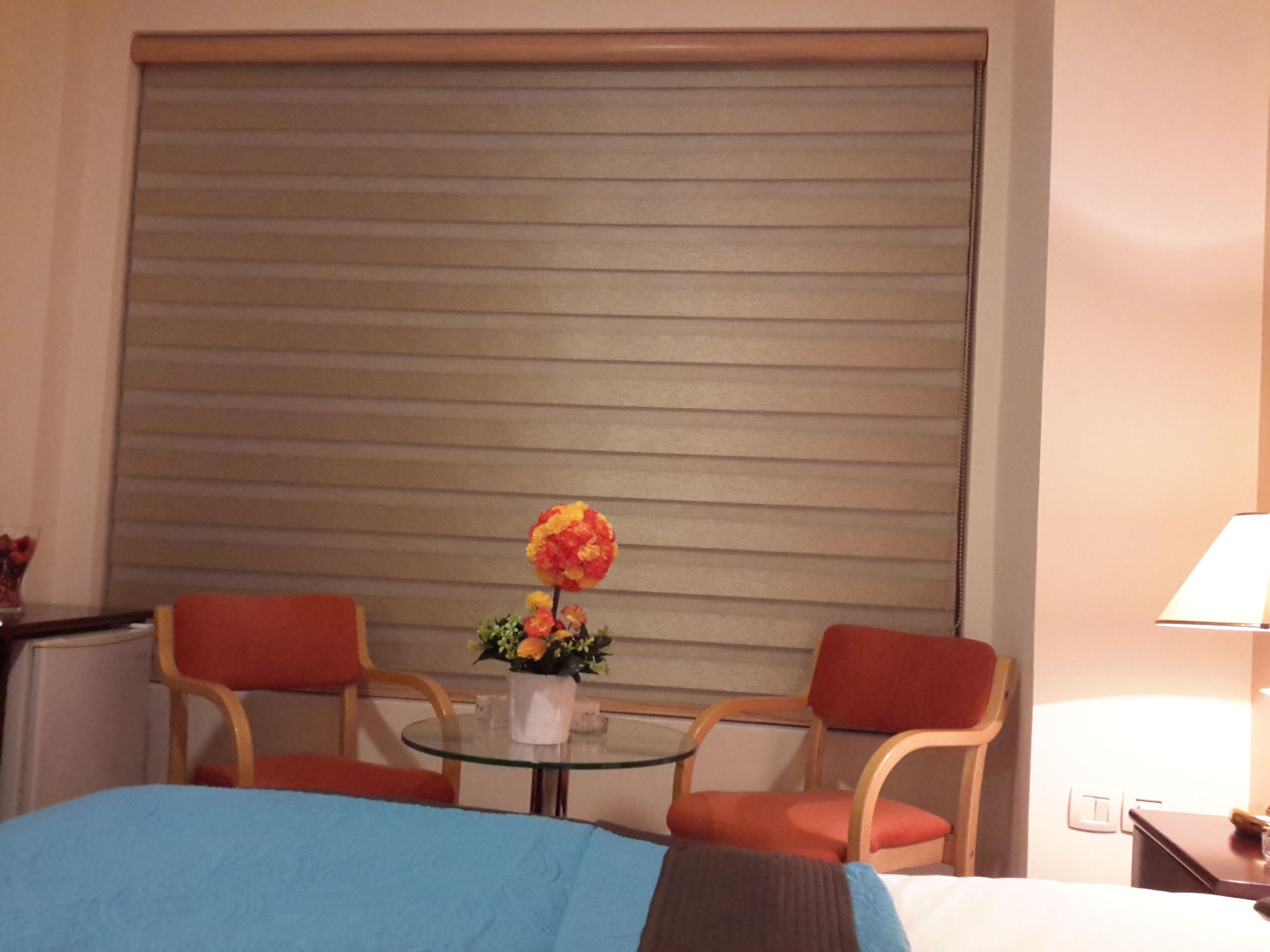 Une chambre d’hôtel avec un lit, une petite table sur laquelle est posé un vase de fleurs, deux chaises et une grande fenêtre fermée par un store.