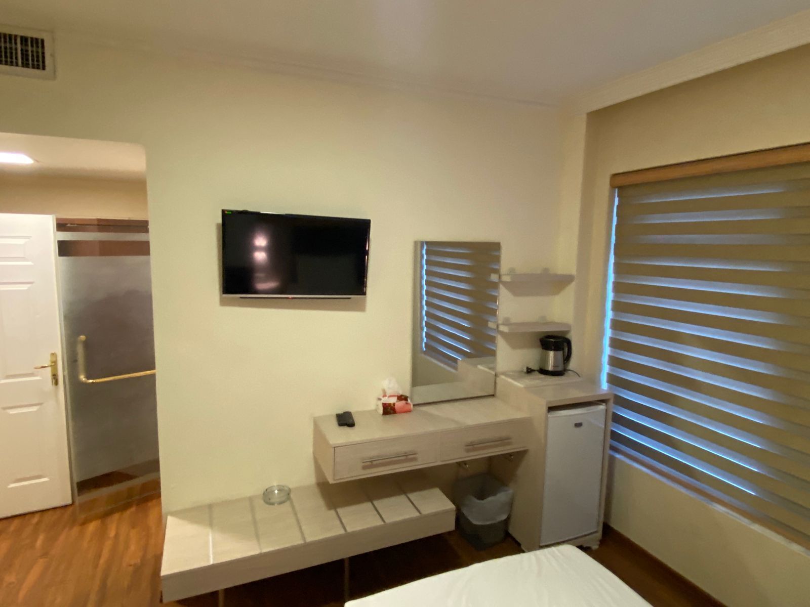 Habitació d'hotel amb un llit, una televisió penjada a la paret, un refrigerador petit, una cafetera i una finestra amb persianes.