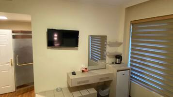 Habitació d'hotel amb un llit, una televisió penjada a la paret, un refrigerador petit, una cafetera i una finestra amb persianes.